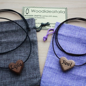 May include: Two necklaces with wooden heart pendants, one engraved with "Layla" and the other with "Feel". The pendants are on black cords, presented in gray and purple drawstring bags. The image includes a business card.
