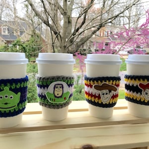 May include: Four white coffee cups with crocheted sleeves featuring different Toy Story characters. The sleeves are striped in various colours, including green, blue, purple, grey, white, red, yellow, and black. Each sleeve has a different character embroidered on it: Buzz Lightyear, Woody, Jessie, and Alien.
