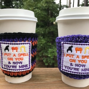 May include: Two crocheted coffee cup sleeves with a white patch featuring the text "I put a spell on you & now you're mine" and three silhouettes of the Sanderson sisters from the movie Hocus Pocus. One sleeve is black and purple, the other is purple with glitter.