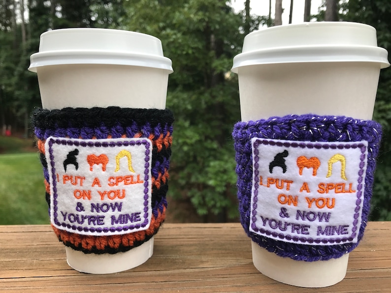 May include: Two crocheted coffee cup sleeves with a white patch featuring the text "I put a spell on you & now you're mine" and three silhouettes of the Sanderson sisters from the movie Hocus Pocus. One sleeve is black and purple, the other is purple with glitter.