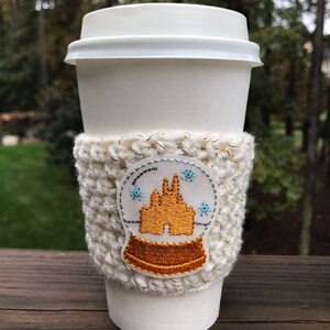 May include: White crocheted coffee cup cozy with a gold embroidered snow globe design featuring a castle. The cozy is on a white coffee cup.