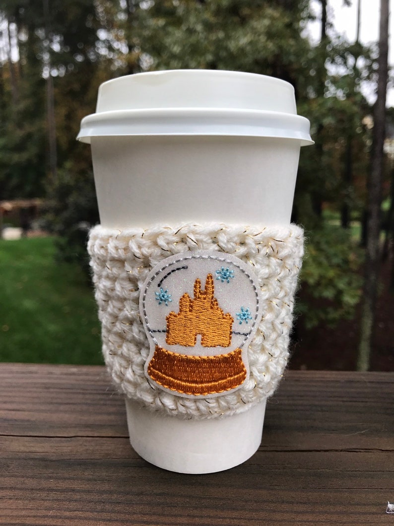 May include: White crocheted coffee cup cozy with a gold embroidered snow globe design featuring a castle. The cozy is on a white coffee cup.