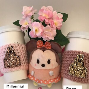May include: Two pink and rose gold coffee cup sleeves with a gold glitter castle design. The sleeves are on white coffee cups. A pink and white plush toy is in the center of the image.