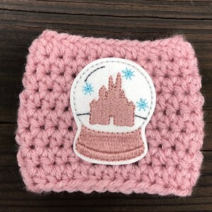 May include: Pink crocheted coffee cup cozy with a white felt snow globe applique featuring a pink embroidered castle and blue snowflakes.