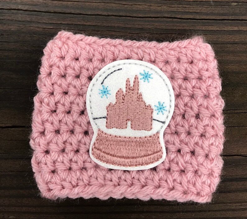 May include: Pink crocheted coffee cup cozy with a white felt snow globe applique featuring a pink embroidered castle and blue snowflakes.