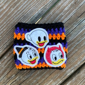 May include: A handmade, crocheted coffee cup sleeve in black, purple, and orange. The sleeve features three appliqued cartoon character faces: a ghost, a witch, and a devil. The characters have yellow beaks and red tongues. The sleeve is designed to fit a standard coffee cup.