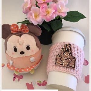 May include: Pink and white crocheted coffee cup cozy with a pink glitter castle and Mickey Mouse design.  The text "Millennial Pink" is at the bottom of the image.