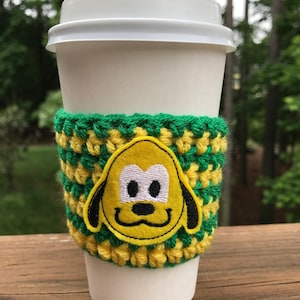 May include: A white coffee cup with a green and yellow crocheted cup cosy featuring a yellow embroidered cartoon dog with black eyes and nose.
