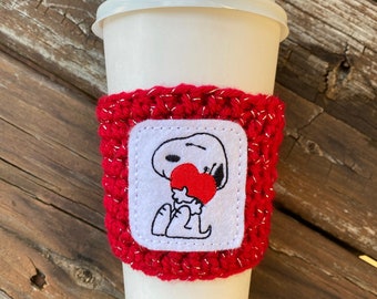 May include: Red crocheted coffee cup cozy with a white embroidered image of Snoopy holding a red heart.