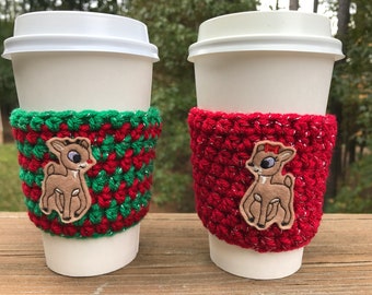 May include: Two white coffee cups with red and green crocheted cup sleeves. Each sleeve has a brown reindeer applique with a red bow.