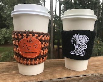 May include: Two coffee cups with crocheted sleeves. One sleeve features a brown and orange pumpkin design, the other a black sleeve with a white embroidered mummy character.