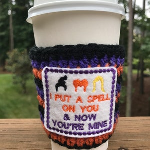 May include: A crocheted coffee cup cozy with a purple, black, and orange striped design. The cozy has a white patch with embroidered black, red, and yellow figures and the text "I put a spell on you & now you're mine".
