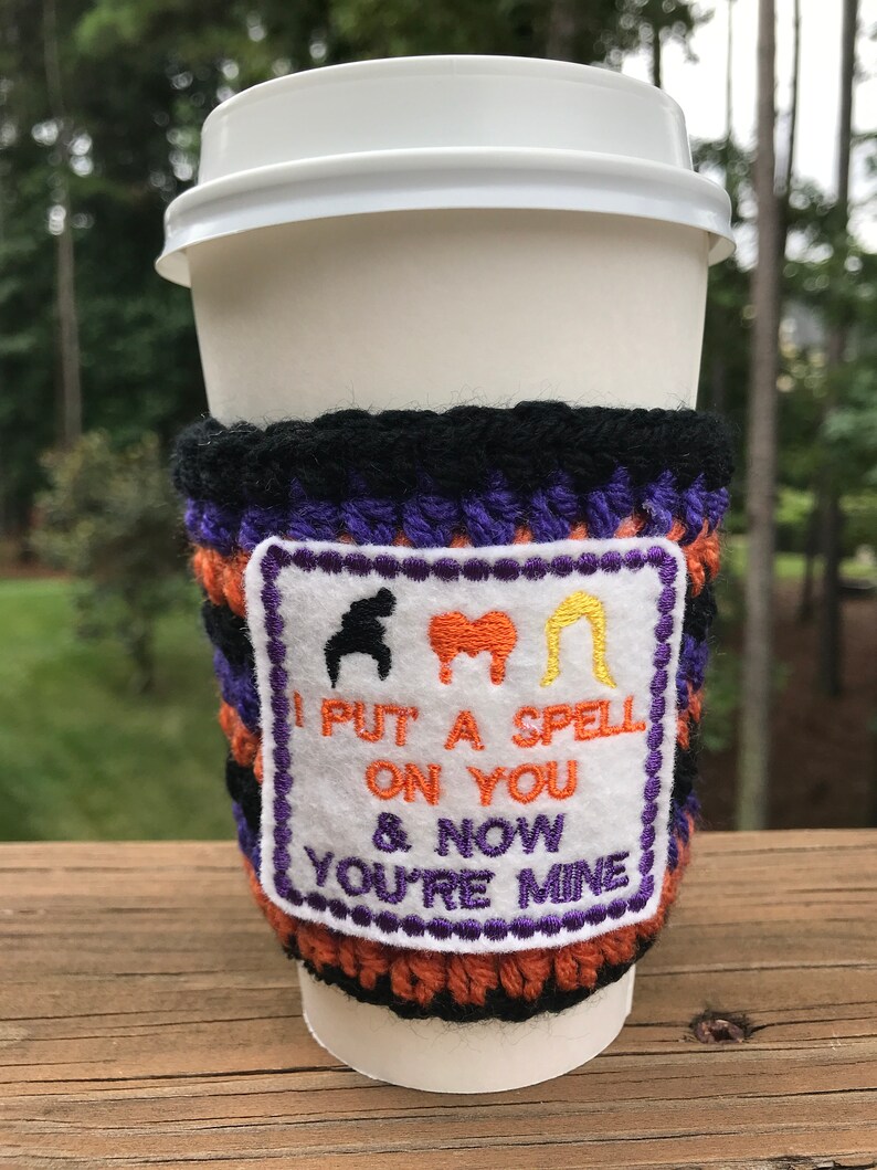 May include: A crocheted coffee cup cozy with a purple, black, and orange striped design. The cozy has a white patch with embroidered black, red, and yellow figures and the text "I put a spell on you & now you're mine".