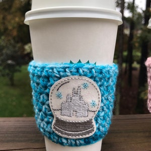 May include: A white to-go coffee cup with a blue crocheted cup cozy. The cozy has a white embroidered snow globe design with a castle inside.