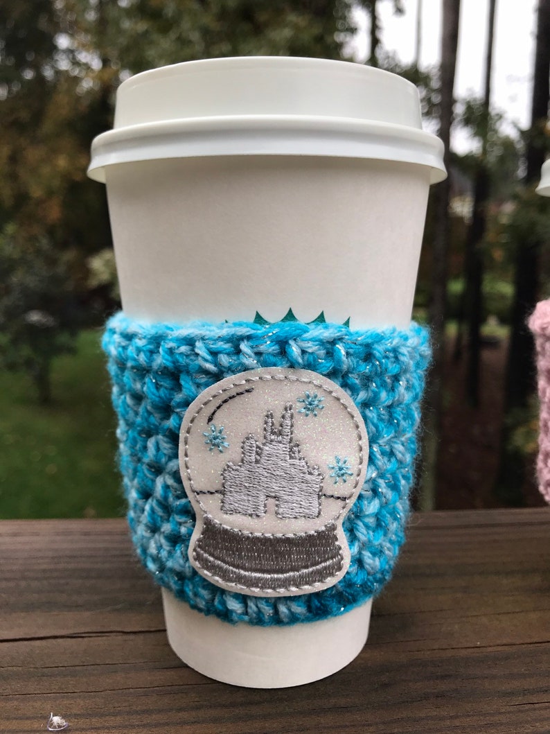 May include: A white to-go coffee cup with a blue crocheted cup cozy. The cozy has a white embroidered snow globe design with a castle inside.