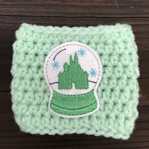 May include: A light green crocheted coffee cup cozy with a white and green embroidered snow globe design featuring a castle.