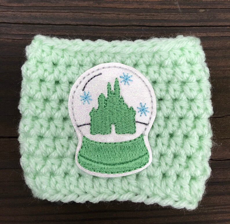 May include: A light green crocheted coffee cup cozy with a white and green embroidered snow globe design featuring a castle.