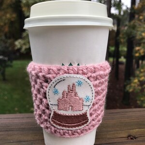 May include: A pink crocheted coffee cup cozy with a white and pink embroidered snow globe design. The snow globe features a pink castle and blue snowflakes.