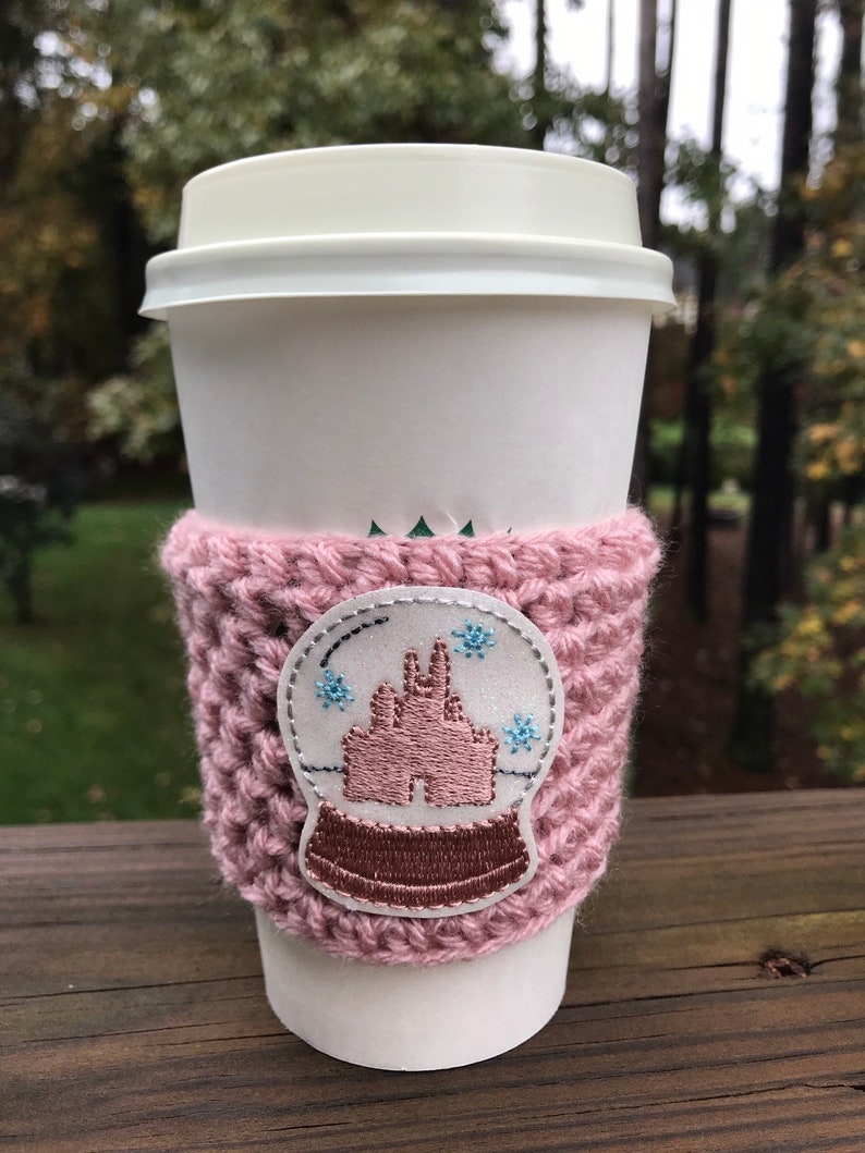May include: A pink crocheted coffee cup cozy with a white and pink embroidered snow globe design. The snow globe features a pink castle and blue snowflakes.