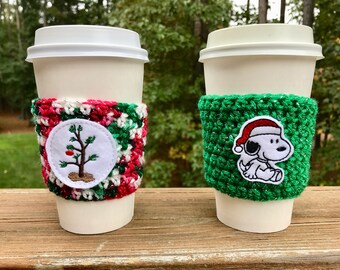 May include: Two white coffee cups with crocheted holiday themed sleeves. One sleeve is green with a white Snoopy wearing a Santa hat. The other sleeve is red, white, and green with a white circle featuring a small green tree with a red ornament.