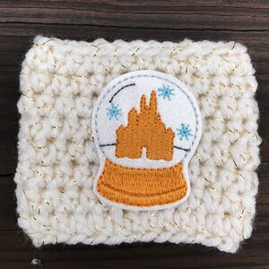 May include: White crocheted coffee cozy with a gold glitter thread. A white and silver snow globe applique with a gold castle inside is centered on the cozy.
