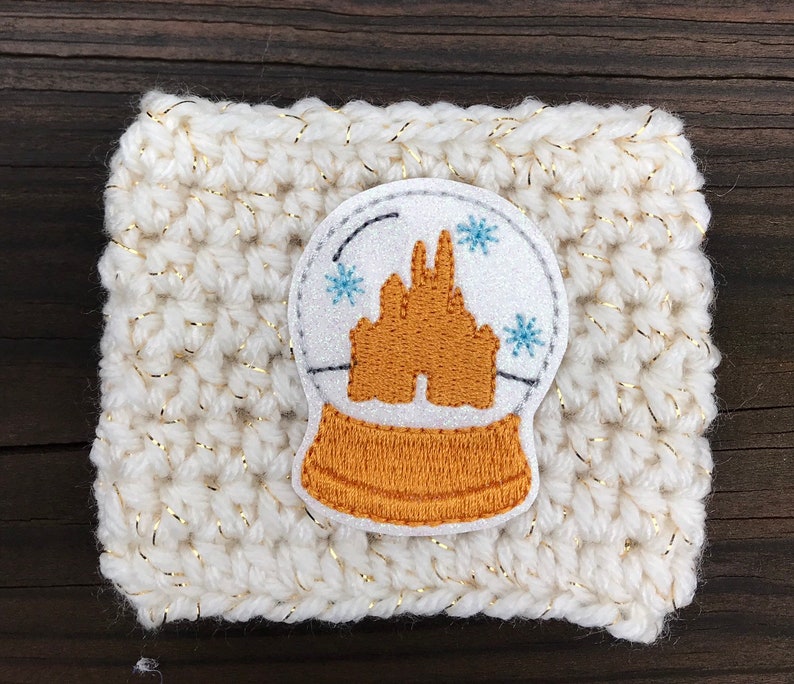 May include: White crocheted coffee cozy with a gold glitter thread. A white and silver snow globe applique with a gold castle inside is centered on the cozy.