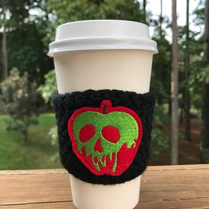 Pu&ograve; includere: Un bicchiere di caff&egrave; bianco da asporto con una manica in crochet nero che presenta una mela rossa ricamata con del veleno verde che cola.
