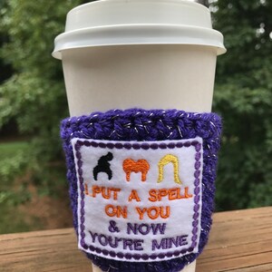 May include: Purple crocheted coffee cup cozy with a white patch that says "I put a spell on you & now you're mine" with three witch silhouettes above the text.
