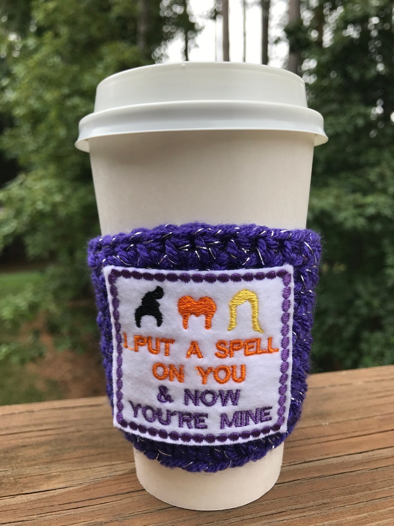 May include: Purple crocheted coffee cup cozy with a white patch that says "I put a spell on you & now you're mine" with three witch silhouettes above the text.