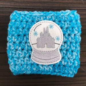 May include: A blue and white crocheted coffee cup cozy with a white and gray embroidered snow globe featuring a gray castle.