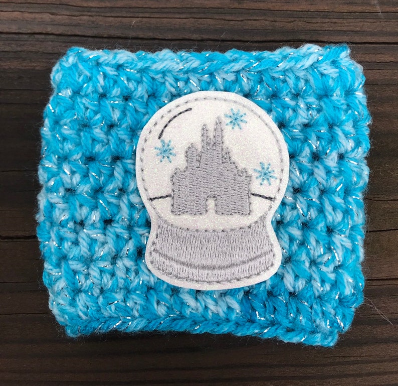 May include: A blue and white crocheted coffee cup cozy with a white and gray embroidered snow globe featuring a gray castle.