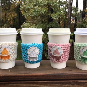 May include: Four white coffee cups with crocheted sleeves featuring a snow globe design with a castle inside. The sleeves are in white, blue, pink, and green.