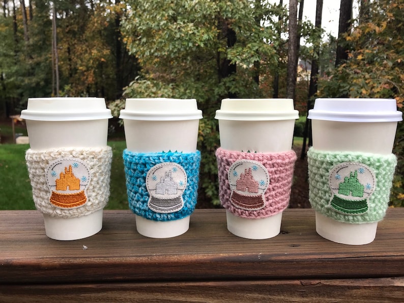 May include: Four white coffee cups with crocheted sleeves featuring a snow globe design with a castle inside. The sleeves are in white, blue, pink, and green.