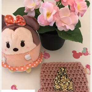 May include: A pink crocheted coffee cup cozy with a gold glitter castle design. The text "Rose Gold" is written below the cozy.