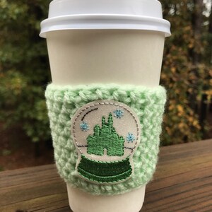 May include: A white coffee cup with a green crocheted cozy. The cozy has a white snow globe design with a green castle inside. The snow globe has blue snowflakes.