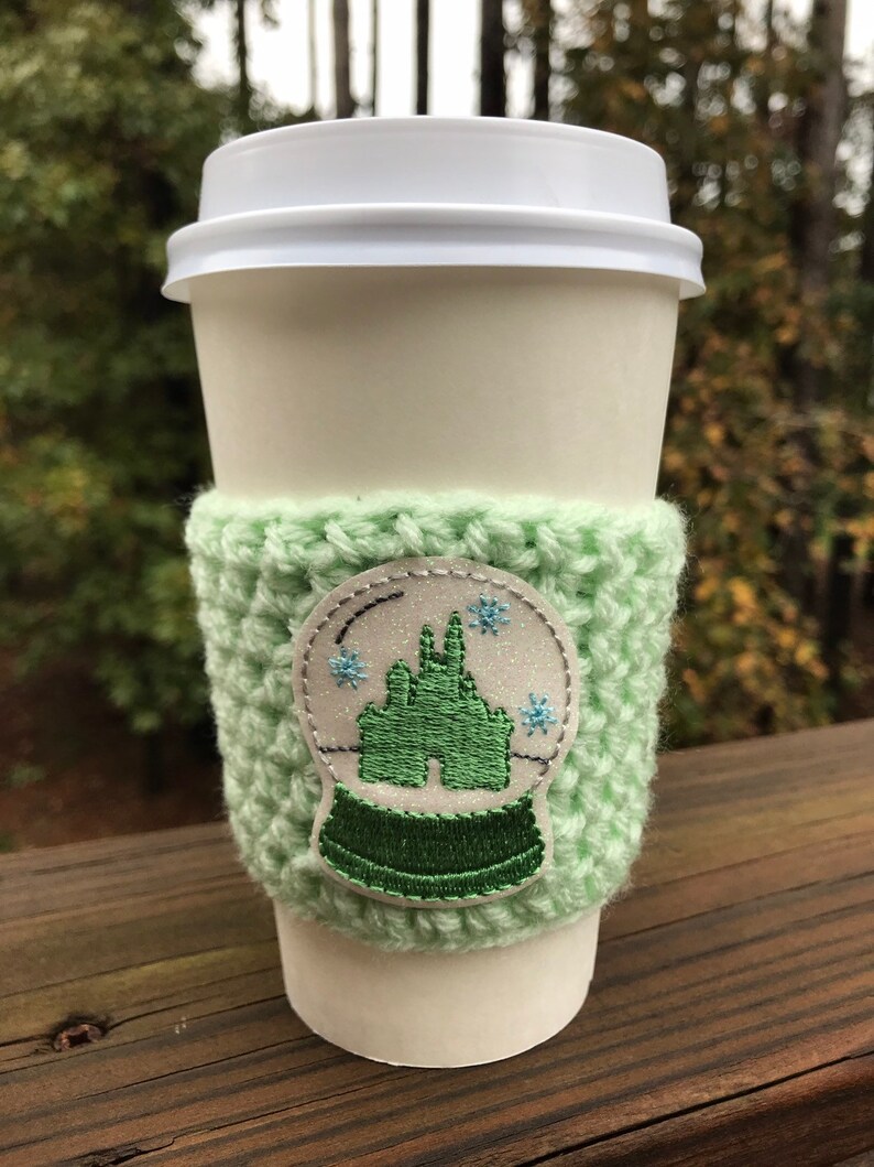 May include: A white coffee cup with a green crocheted cozy. The cozy has a white snow globe design with a green castle inside. The snow globe has blue snowflakes.