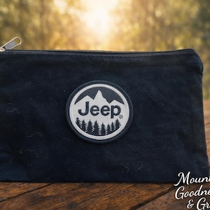 May include: A black canvas zippered pouch with a circular patch featuring a mountain and pine tree design, with the word "Jeep" above the trees. The pouch is on a wooden surface.