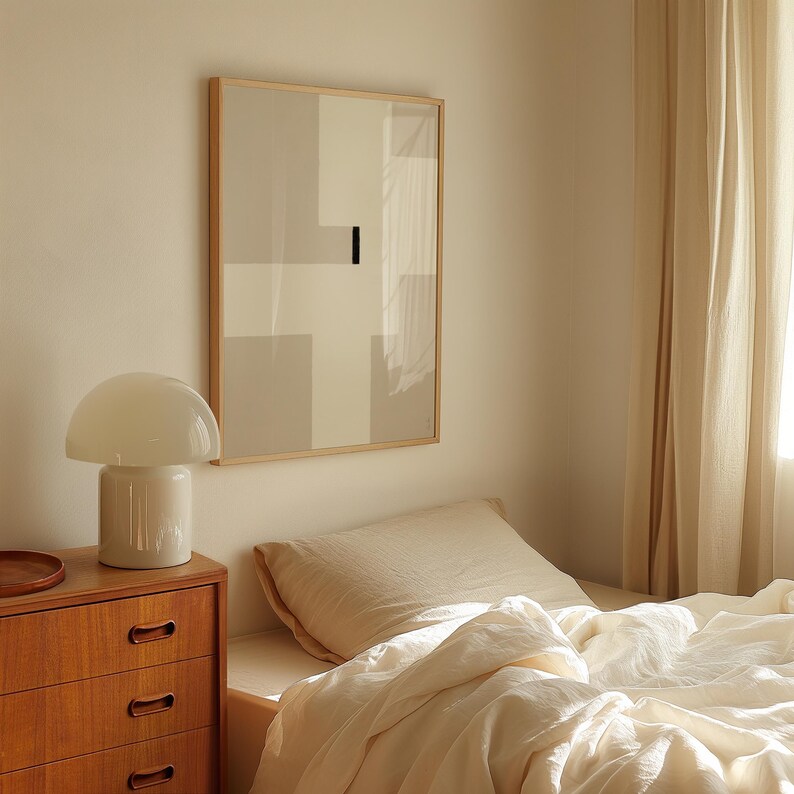 May include: A white bedside lamp with a mushroom-shaped shade sits on a wooden dresser with drawers. A framed abstract art print with geometric shapes in shades of gray hangs on the wall behind the lamp. A white bed with a white duvet and a beige pillow is in the background.