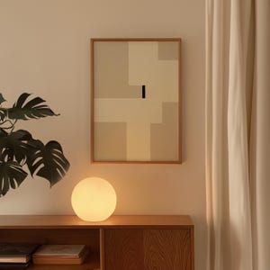 May include: A white globe lamp with a warm glow sits on a wooden table next to a framed abstract painting with beige and white geometric shapes. The painting is hanging on a light beige wall.