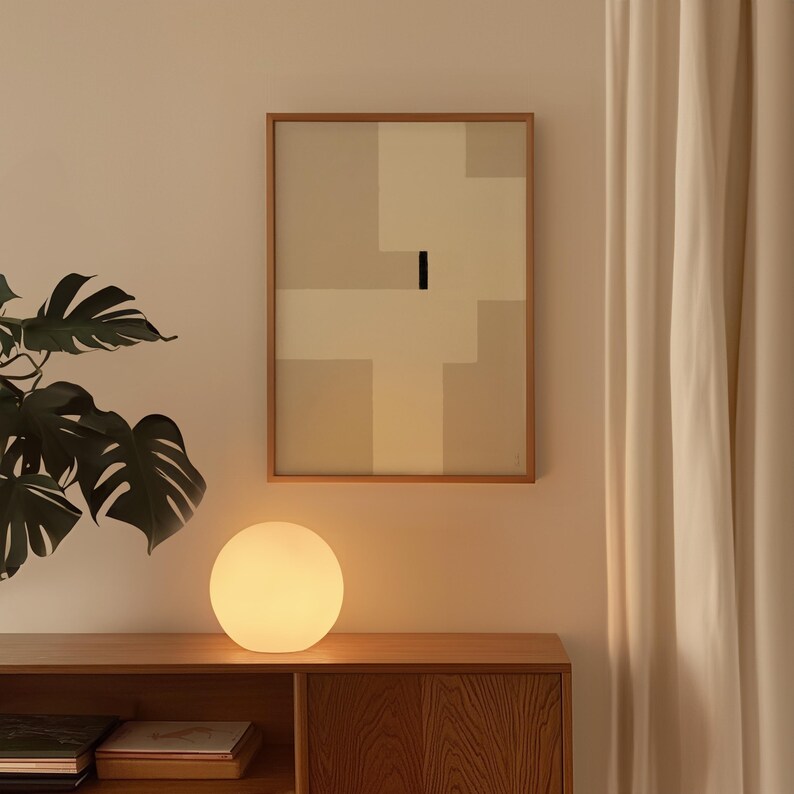 May include: A white globe lamp with a warm glow sits on a wooden table next to a framed abstract painting with beige and white geometric shapes. The painting is hanging on a light beige wall.