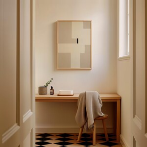 May include: A light wood desk with a white towel draped over a wooden stool. A small potted plant, a bottle of hand soap, and a white rectangular object are on the desk. A framed abstract painting with beige and white geometric shapes hangs on the wall behind the desk. The floor is black and white checkered tile.