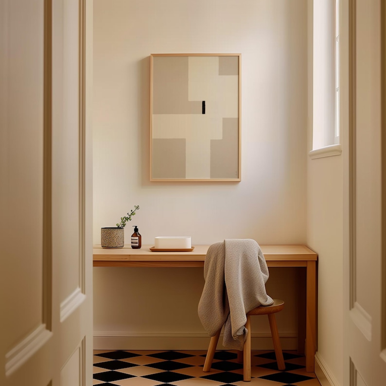 May include: A light wood desk with a white towel draped over a wooden stool. A small potted plant, a bottle of hand soap, and a white rectangular object are on the desk. A framed abstract painting with beige and white geometric shapes hangs on the wall behind the desk. The floor is black and white checkered tile.