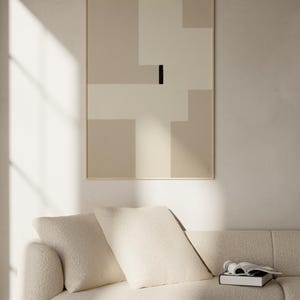 May include: A white couch with two pillows and a book on the seat. A framed abstract art print with beige and white geometric shapes and a black line hangs on the wall behind the couch.