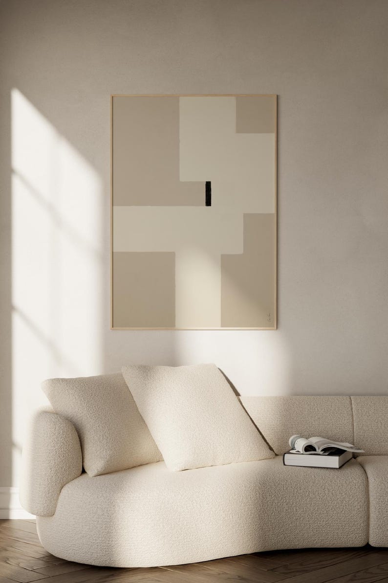 May include: A white couch with two pillows and a book on the seat. A framed abstract art print with beige and white geometric shapes and a black line hangs on the wall behind the couch.