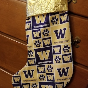 Puede incluir: Una media de Navidad dorada y morada con un patrón repetido de la letra "W" y una huella de pata. La media tiene el texto "WASHINGTON®" en ella.