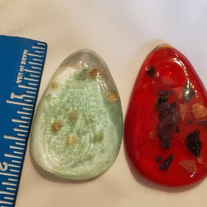 May include: Two teardrop-shaped pendants. One is light green with gold flecks, the other is red with dark and gold inclusions. Both have a hole at the top for hanging. A blue ruler with inch markings is on the left.