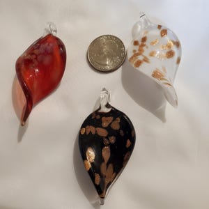 May include: Three glass ornaments in red, black, and white with gold accents. The ornaments have a teardrop shape and are displayed with a quarter for size comparison. The red ornament is swirled, the black has gold speckles, and the white has gold spots.
