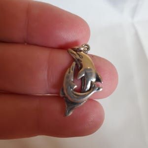 May include: A silver-toned pendant featuring two dolphins intertwined. The dolphins are depicted in a stylized, three-dimensional design, with smooth, curved bodies and a small loop at the top for a chain. The pendant is held against a neutral background.