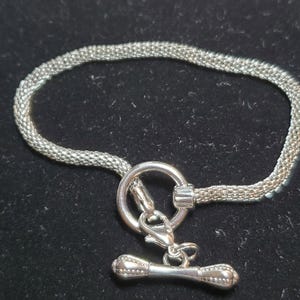 May include: A silver-colored bracelet with a mesh chain design. The bracelet features a toggle clasp with a round ring and a bar closure. The bracelet is displayed on a dark surface.