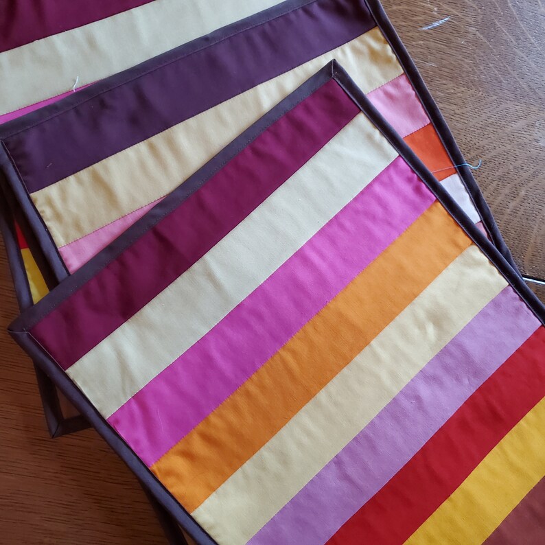 May include: Set of three striped fabric placemats in shades of red, orange, yellow, pink, purple, and brown. The placemats have a dark brown border.